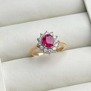 May include: A gold ring featuring a central oval-cut red ruby surrounded by a halo of small, clear, round-cut white diamonds. The ruby is set in a prong setting, and the band is made of polished yellow gold, creating a classic, multi-stone design.