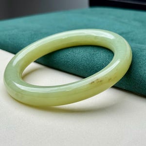 Può includere: Un braccialetto bangle in giada verde pallido. La superficie lucida riflette la luce, mettendo in risalto le variazioni naturali di colore. Il braccialetto è di forma ovale e sembra essere un pezzo unico di giada. Lo sfondo è un tessuto verde acqua a contrasto.