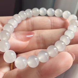 K&ouml;nnte beinhalten: Ein elastisches Armband aus glatten, runden, durchscheinenden wei&szlig;en Perlen mit einem dezenten opalisierenden Schimmer. Die Perlen sind einheitlich gro&szlig; und zu einer kreisf&ouml;rmigen Schlaufe aufgereiht, wobei sie das Licht auf ihren polierten Oberfl&auml;chen reflektieren.