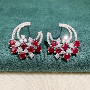 May include: A pair of silver-toned earrings with a crescent design. Each earring features a cluster of round, deep red gemstones and smaller white gemstones. The earrings are set against a green background.