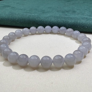 May include: A circular bracelet composed of smooth, polished, light lavender-grey spherical beads. The beads are uniform in size and shape, strung together to form a continuous loop. The surface of each bead reflects light, showing a subtle sheen.