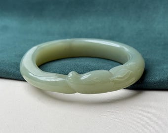 May include: A pale green jade bangle bracelet. The bracelet features a carved dragon head design on one side. The smooth, polished surface reflects light, highlighting the natural variations in the stone. The bracelet is displayed on a white and teal background.