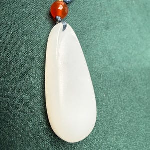May include: A white, teardrop-shaped pendant hangs from a blue cord, accented by a small, orange bead. The pendant has a smooth, polished surface. The background is a textured, dark green fabric.