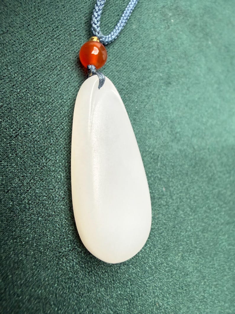 May include: A white, teardrop-shaped pendant hangs from a blue cord, accented by a small, orange bead. The pendant has a smooth, polished surface. The background is a textured, dark green fabric.