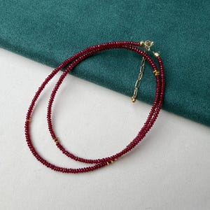 May include: A delicate necklace featuring small, deep red beads strung together. The necklace has a gold-colored clasp and small gold accents. The necklace is displayed on a white surface with a green fabric background.