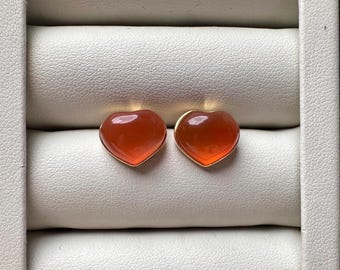 Puede incluir: Un par de pendientes de botón con piedras de cornalina naranja en forma de corazón, lisas y pulidas, engastadas en monturas de metal dorado. Los pendientes tienen un aspecto brillante y translúcido y se muestran uno al lado del otro.