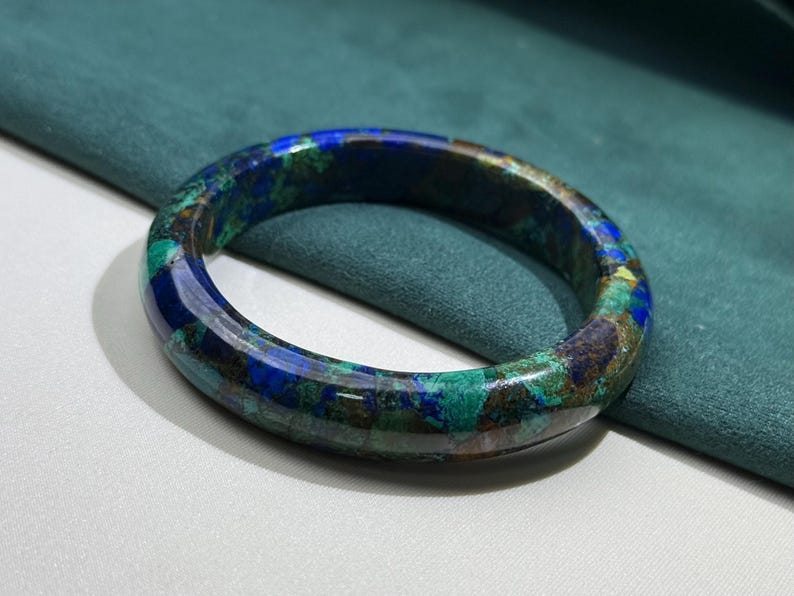 May include: A polished, round bracelet with a marbled appearance. The bracelet features a blend of blue, green, and brown hues. The gemstone bracelet is a piece of jewelry.