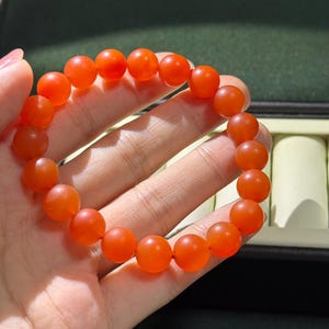 May include: A stretch bracelet composed of numerous smooth, round, translucent orange beads. The beads are uniform in size and shape, creating a continuous circular loop. The material has a soft, matte-like finish that catches the light.
