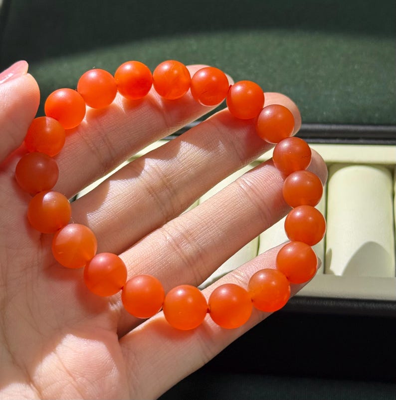 May include: A stretch bracelet composed of numerous smooth, round, translucent orange beads. The beads are uniform in size and shape, creating a continuous circular loop. The material has a soft, matte-like finish that catches the light.