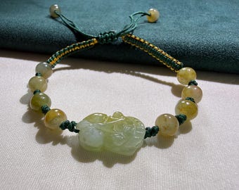 Op de afbeelding: Een armband met een centrale, lichtgroene, jade-achtige hanger in de vorm van een mythisch wezen, geflankeerd door ronde, doorschijnende geelgroene kralen aan een donkergroen gevlochten koord met een verstelbare schuifknoopsluiting.