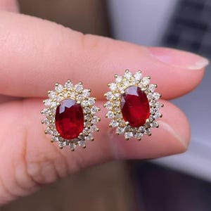 May include: A pair of gold stud earrings, each featuring a vibrant oval-cut ruby surrounded by a halo of small, sparkling diamonds. The earrings are set in a gold metal setting, creating a luxurious and eye-catching design.