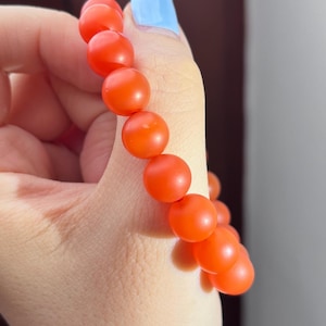 May include: A bracelet composed of multiple smooth, round, vibrant orange beads. The beads have a matte finish and are strung together in a single strand, creating a uniform circular shape that fits around the wrist.
