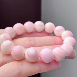 May include: A stretch bracelet composed of smooth, spherical beads in varying shades of soft pastel pink and white. Each bead features a subtle, concentric circular pattern, creating a layered appearance throughout the circular jewelry piece.