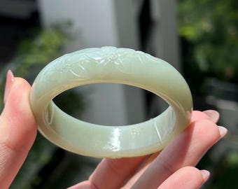 May include: A pale green jade bangle bracelet. The bracelet features a floral pattern carved around the exterior. The smooth, polished surface reflects light, highlighting the intricate design. A piece of jewelry.