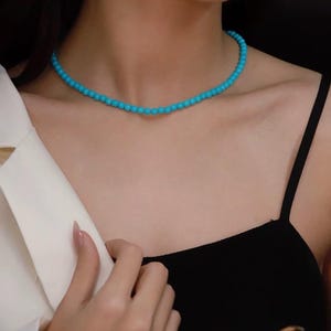 Pode incluir: Um delicado colar de contas turquesa. O colar é composto por pequenas contas redondas azuis claras, enfiadas para formar um colar estilo gargantilha. O colar é usado com um top preto e um blazer branco.