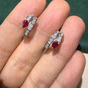 May include: A pair of silver-toned earrings in a snake design, featuring a pear-shaped red gemstone at the head and small, clear gemstones along the body. The earrings are displayed on a person's fingers.