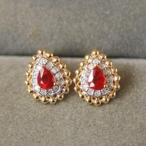 May include: A pair of gold stud earrings featuring a central pear-shaped red gemstone surrounded by a halo of small white stones. The outer edge of each earring has a textured, beaded gold border, creating a classic and detailed appearance.