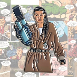 May include: An enamel pin depicting a woman with long black hair holding a blue and silver weapon. She is wearing a brown jacket with a white shirt underneath and a black belt. The pin features a white circle with a blue and silver symbol in the centre. The background is a comic book panel.