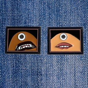 May include: Two enamel pins depicting a cartoon character with a single eye and a mouth. The character on the left has a toothy grin, while the character on the right has a closed-mouth smile. Both pins are set against a blue denim background.