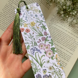 Op de afbeelding: Een witte bladwijzer met een bloemenpatroon met verschillende bloemen, waaronder blauwe bellen, klaprozen en madeliefjes. De bladwijzer heeft een groene kwast.