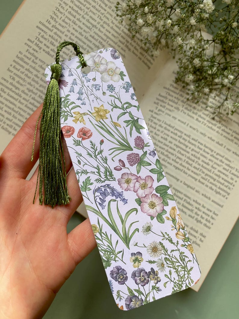 Op de afbeelding: Een witte bladwijzer met een bloemenpatroon met verschillende bloemen, waaronder blauwe bellen, klaprozen en madeliefjes. De bladwijzer heeft een groene kwast.