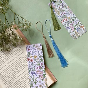 Op de afbeelding: Twee bloemen-bladwijzers met kwastjes. De bladwijzers hebben een witte achtergrond met een verscheidenheid aan kleurrijke bloemen en groen. De kwastjes zijn groen, blauw en beige.