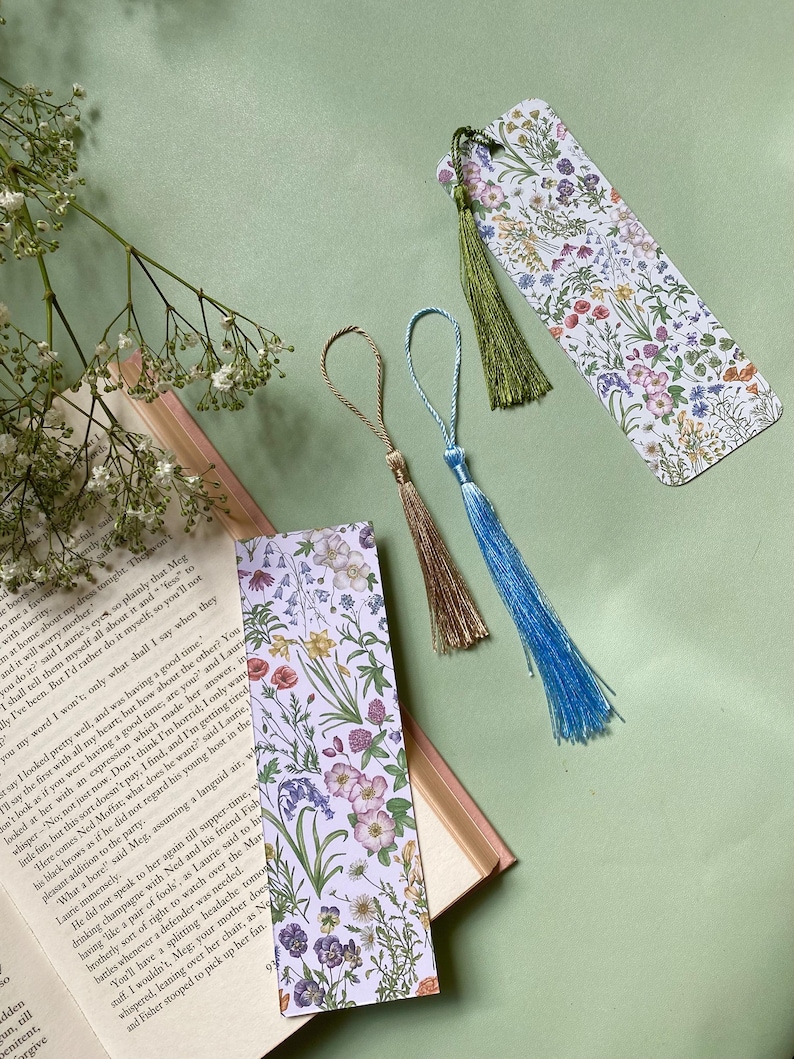 Op de afbeelding: Twee bloemen-bladwijzers met kwastjes. De bladwijzers hebben een witte achtergrond met een verscheidenheid aan kleurrijke bloemen en groen. De kwastjes zijn groen, blauw en beige.