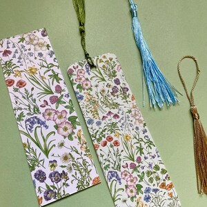 Op de afbeelding: Twee boekenlegger met bloemenmotief en groene, blauwe en gouden kwastjes. De boekenleggers zijn wit met een verscheidenheid aan kleurrijke bloemen en groen.