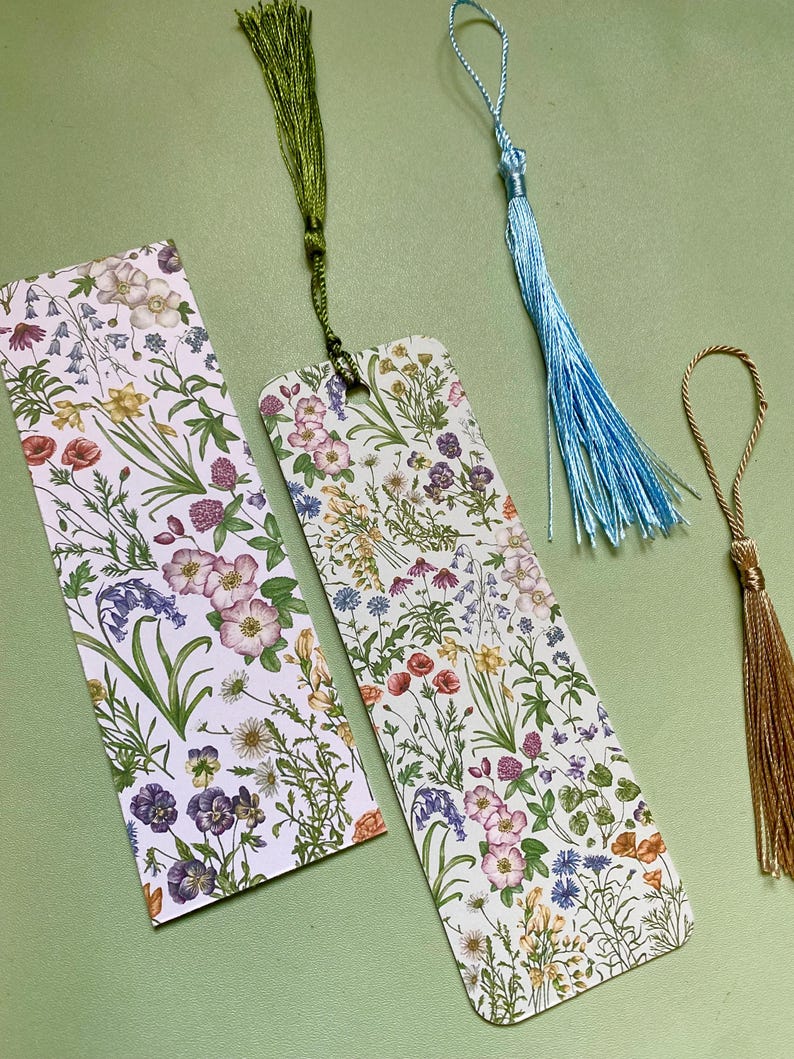 Op de afbeelding: Twee boekenlegger met bloemenmotief en groene, blauwe en gouden kwastjes. De boekenleggers zijn wit met een verscheidenheid aan kleurrijke bloemen en groen.