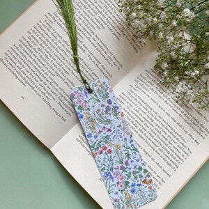 Op de afbeelding: Een witte bladwijzer met een bloemenpatroon met verschillende gekleurde bloemen en groene bladeren. De bladwijzer heeft een kwast aan de bovenkant.