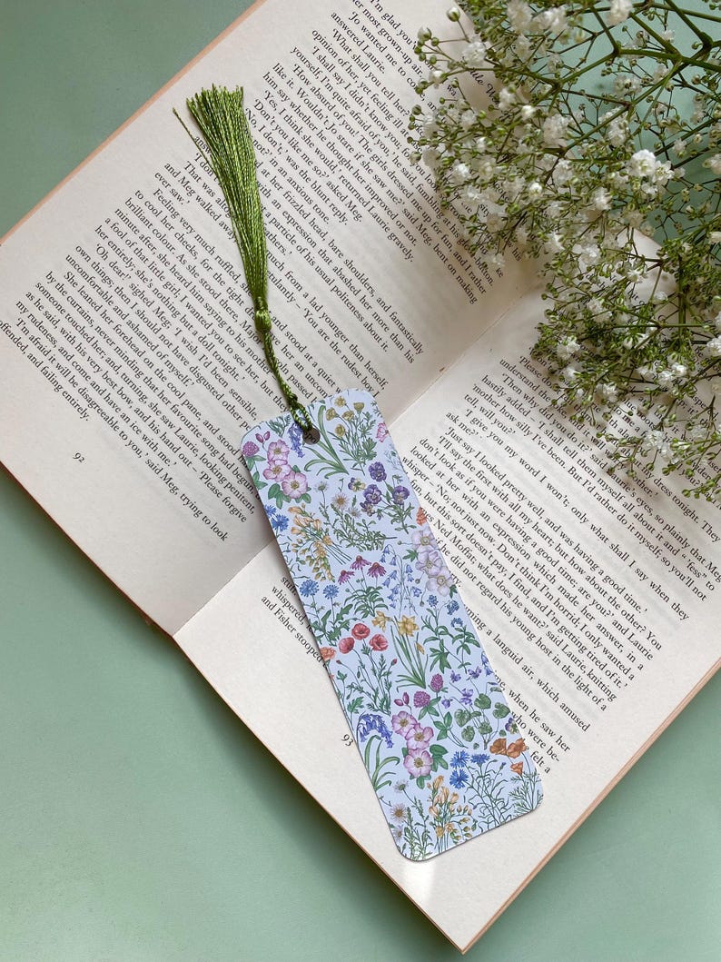 Op de afbeelding: Een witte bladwijzer met een bloemenpatroon met verschillende gekleurde bloemen en groene bladeren. De bladwijzer heeft een kwast aan de bovenkant.