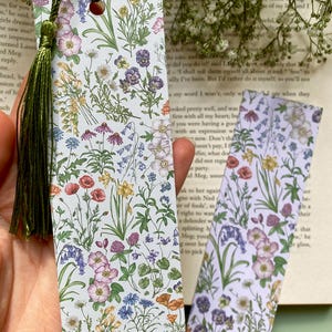 Op de afbeelding: Twee bloemen-bladwijzers met een groene kwast op de linker bladwijzer. De bladwijzers hebben een witte achtergrond met een verscheidenheid aan kleurrijke bloemen en planten.