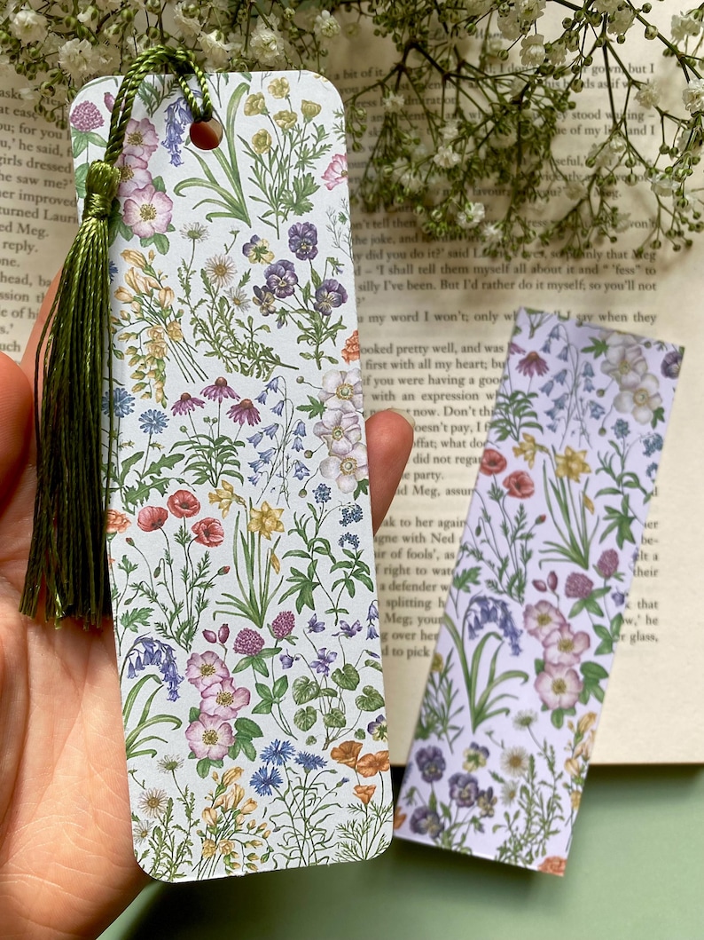 Op de afbeelding: Twee bloemen-bladwijzers met een groene kwast op de linker bladwijzer. De bladwijzers hebben een witte achtergrond met een verscheidenheid aan kleurrijke bloemen en planten.