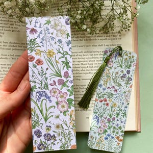 Op de afbeelding: Twee bloemen-bladwijzers met een witte achtergrond en kleurrijke bloemen. De bladwijzers zijn rechthoekig en hebben een groene kwast aan de rechterkant.