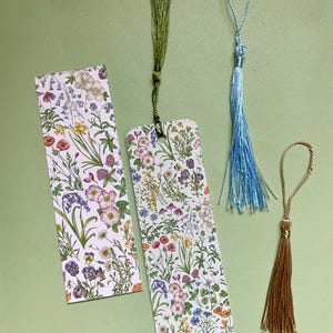Op de afbeelding: Drie bloemmotief-bladwijzerontwerpen met verschillende gekleurde kwastjes. De bladwijzers hebben een witte achtergrond met een herhalend patroon van kleurrijke bloemen en groen. De kwastjes zijn groen, blauw en goud.