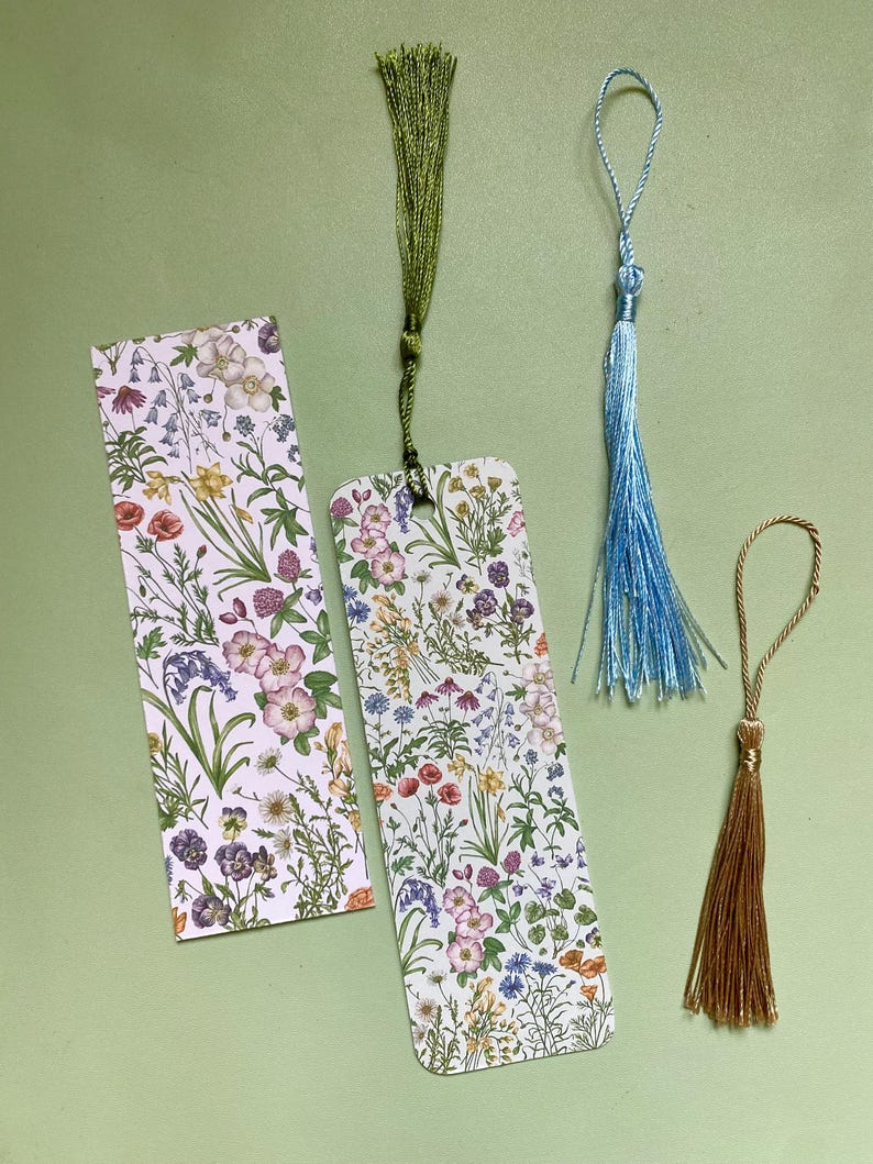 Op de afbeelding: Drie bloemmotief-bladwijzerontwerpen met verschillende gekleurde kwastjes. De bladwijzers hebben een witte achtergrond met een herhalend patroon van kleurrijke bloemen en groen. De kwastjes zijn groen, blauw en goud.