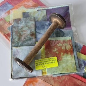 May include: A pack of ice dyed fabric scraps in various colours, including blue, green, yellow, orange, and red. The fabric scraps are arranged in a clear plastic bag with a wooden spool and a yellow label that reads "Colour Creativity Pack At least 50g of ice dyed fabric (FQ equivalent) £9.9".