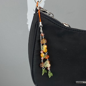 May include: A black handbag with a silver chain strap and a charm attached to the strap. The charm is made of orange string and features small green leaves and clear and amber colored beads.