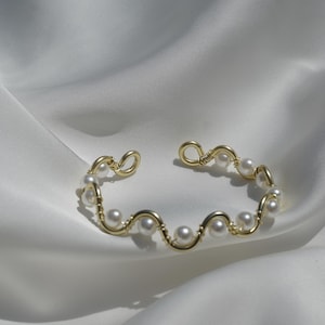 Può includere: Un bracciale a fascia in filo metallico dorato caratterizzato da un design ondulato continuo. Il filo è decorato con piccole perle bianche rotonde e artificiali distanziate uniformemente lungo le curve. Il bracciale ha una struttura aperta.