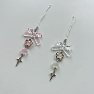 Könnte beinhalten: Zwei transparente Handy-Anhänger mit Schleifen-, Blumen- und Sterndesign. Ein Anhänger hat eine rosa Schleife und Blume, der andere hat eine transparente Schleife und Blume. Beide Anhänger haben einen silberfarbenen Sternanhänger unten.