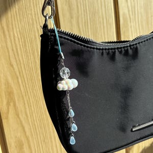 Könnte beinhalten: Eine schwarze Handtasche mit silbernem Reißverschluss und einem dekorativen Anhänger. Der Anhänger zeigt eine weiße Wolke mit rosa Akzenten, eine klare Perle und drei blaue tropfenförmige Perlen. Die Tasche hängt an einer silbernen Kette.