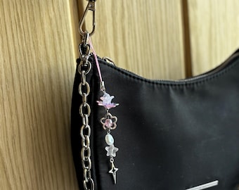 May include: Black shoulder bag with a silver chain strap and a decorative charm. The charm features pink and white flower-shaped beads, a pearl-like bead, and a silver star pendant. The bag is made of a smooth, matte material.