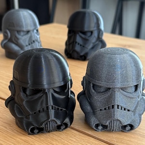 Puede incluir: Varios modelos de cascos de Stormtrooper impresos en 3D, en negro y gris, están dispuestos sobre una superficie de madera. Los cascos presentan detalles icónicos, mostrando el diseño del personaje de Star Wars. Son objetos de colección.
