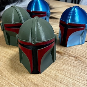 Peut inclure: Quatre casques imprimés en 3D, rappelant un design Mandalorien. Les casques sont dans des tons de vert, de bleu et de gris, avec des accents rouges et noirs. Ils sont disposés sur une surface en bois.
