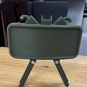 Könnte beinhalten: Ein grünes Schild aus Kunststoff mit der Aufschrift "FRONT TOWARD ENEMY". Das Schild ist auf einem Stativ montiert.