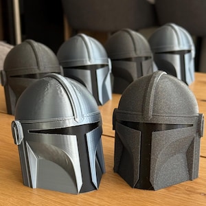 May include: Several Mandalorian helmets in various shades of gray and black are arranged on a wooden surface. The helmets have a distinctive T-shaped visor and are likely 3D-printed, showcasing a detailed design.