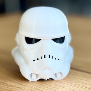 Op de afbeelding: Een witte Stormtrooper-helm, een personage uit de Star Wars-franchise, wordt weergegeven op een houten oppervlak. De helm heeft zwarte ooglenzen en een gedetailleerd gezichtsontwerp, inclusief een reeks verticale lijnen onder de mond.