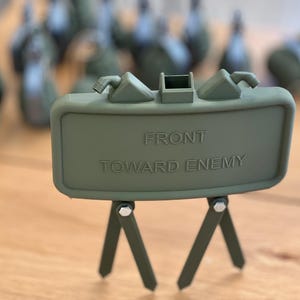 Könnte beinhalten: Ein grünes, rechteckiges Modell einer Landmine mit dem Text "FRONT TOWARD ENEMY". Das Modell wird von drei kleinen Beinen getragen und hat mehrere ähnliche Modelle im Hintergrund verschwommen. Die Objekte befinden sich auf einer Holzoberfläche.