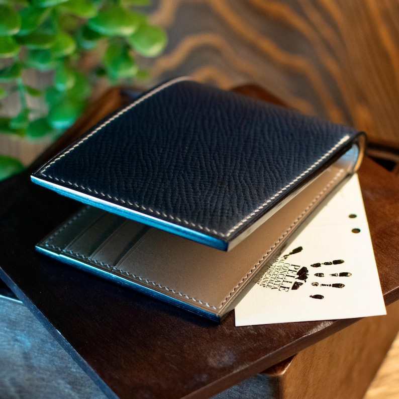 May include: A navy blue leather wallet with white stitching and a light brown interior. The wallet is open, revealing card slots. A small card with a handprint is tucked inside. The wallet is on a wooden surface.