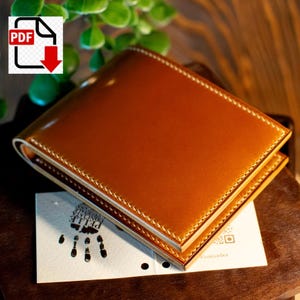May include: A brown leather wallet with white stitching. The wallet is rectangular and appears to be bi-fold. A small card with a handprint and text is visible beneath the wallet. The wallet is sitting on a wooden surface.